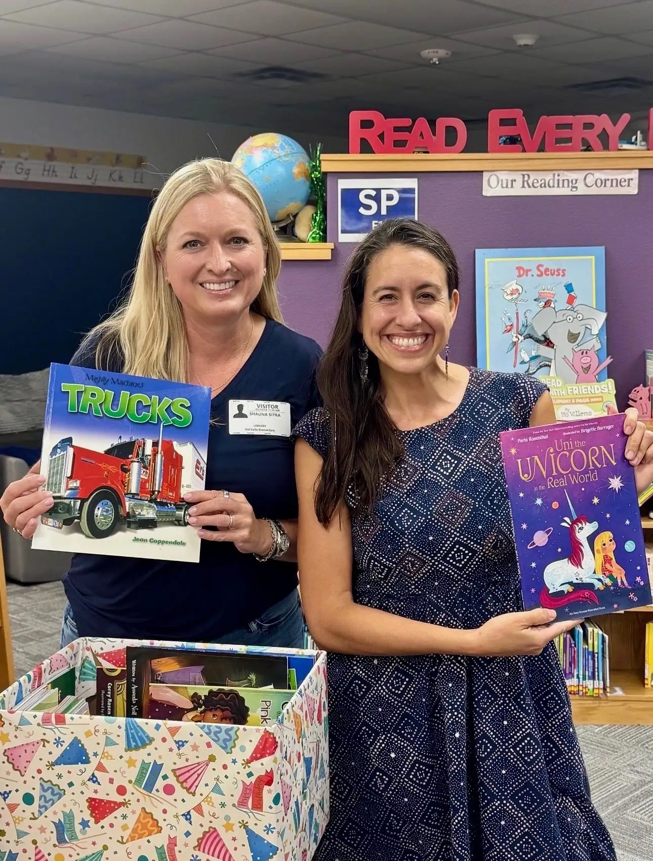 A National Charity League volunteer and a school librarian hold up brand-new books, a truck book and a unicorn picture book, beside a stocked Birthday Box at Del Valle Elementary