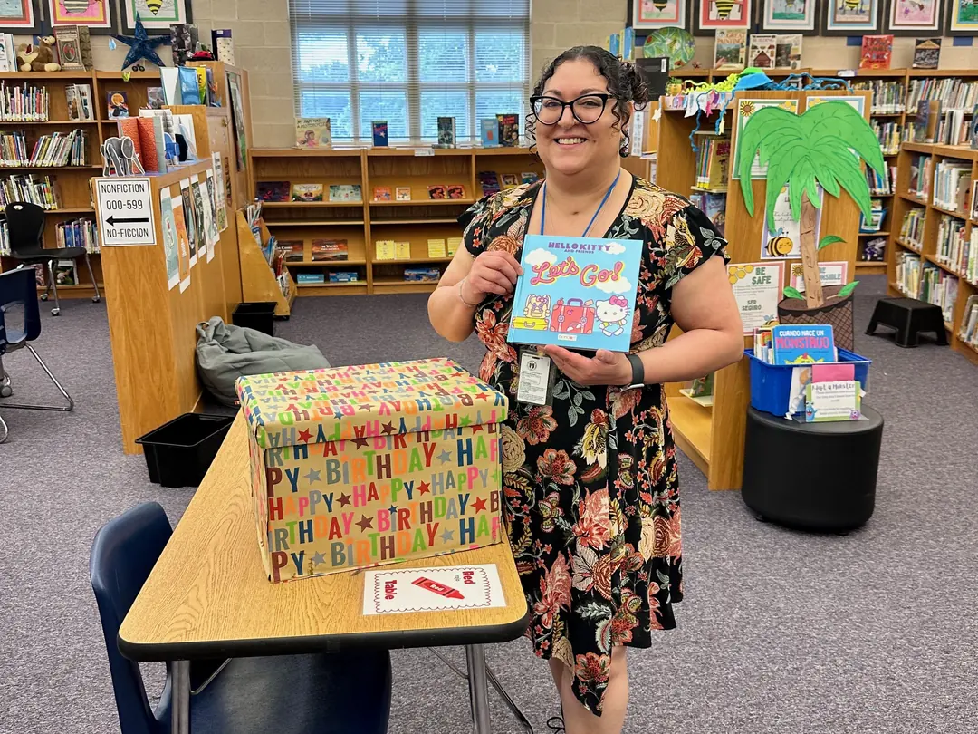 A school librarian smiles while holding a new picture book beside a decorated Birthday Box in the McBee Elementary library