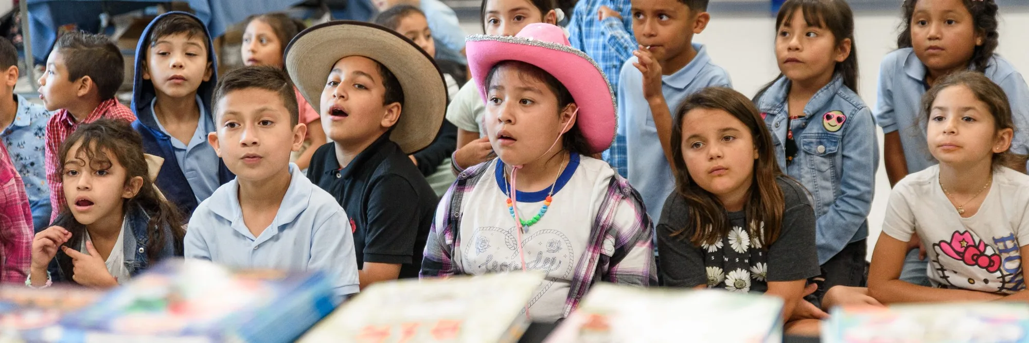 A room of students leans in as their book fair begins, brand-new books stacked on the tables in front of them