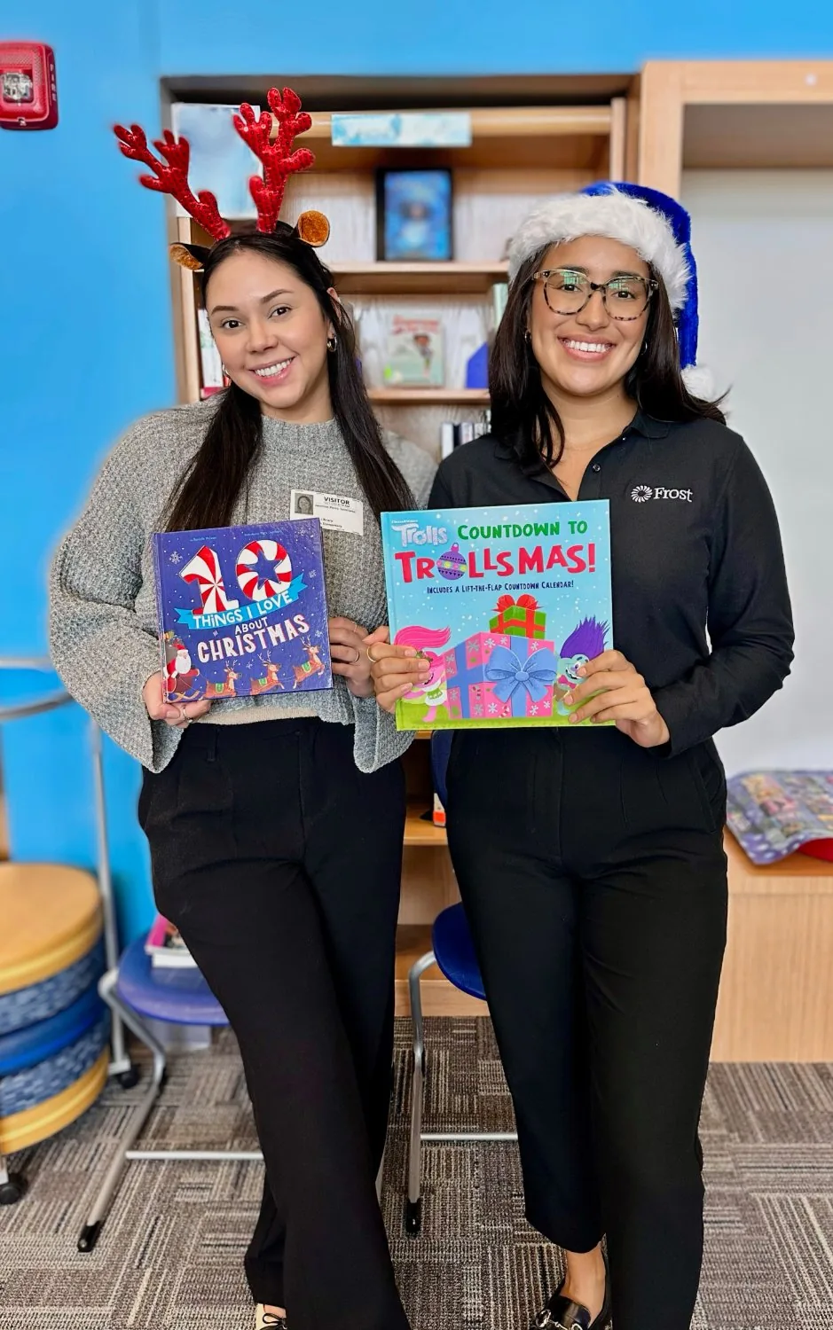 Two Frost Bank employees volunteer at a Book Drive for Kids fair, holding brand-new books in a school library.