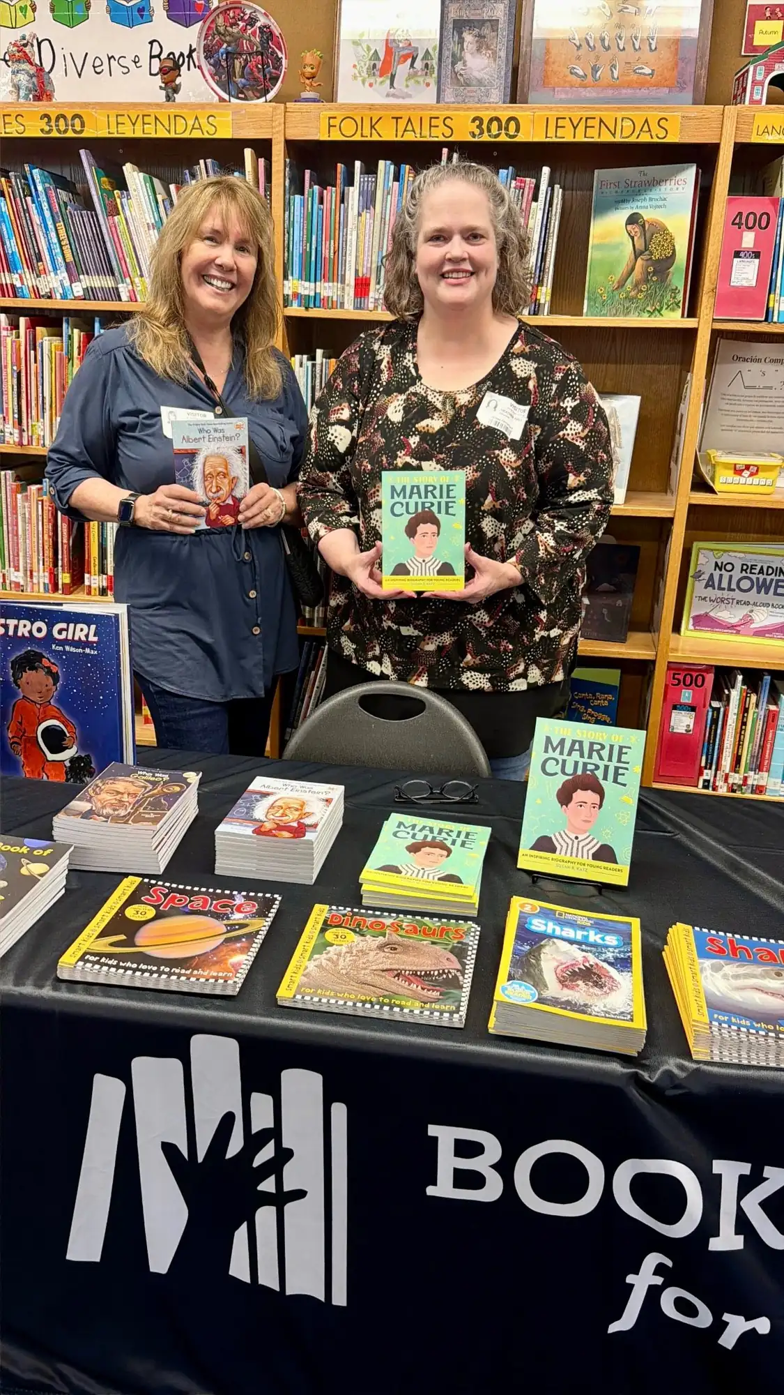 Two volunteers with visitor badges stand behind the Book Drive for Kids table, holding biography readers in a school library