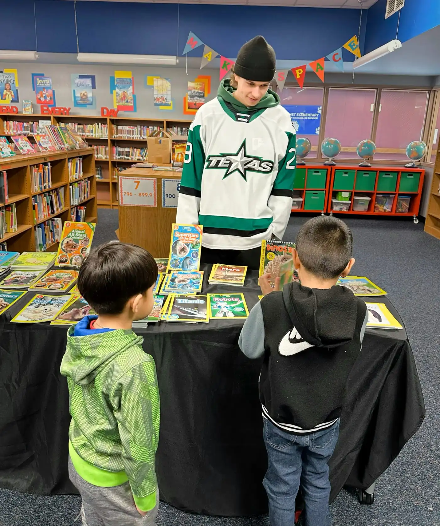A Texas Stars hockey player in his jersey stands behind the book table while two students browse National Geographic readers at Bluebonnet Elementary