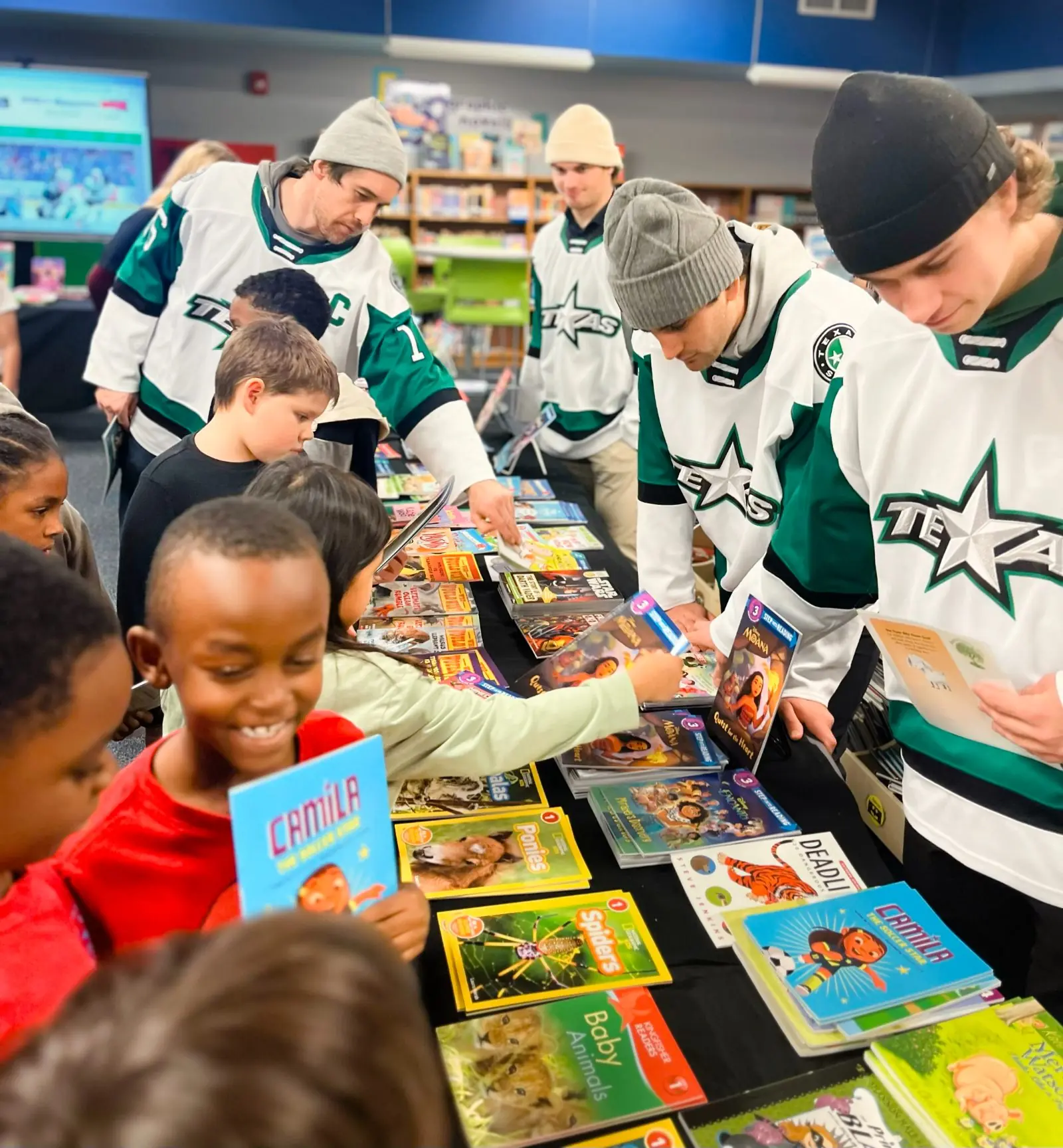 Texas Stars players in jerseys help a crowd of students choosing books at a Bluebonnet Elementary book fair, one girl holding up Camila the Soccer Star