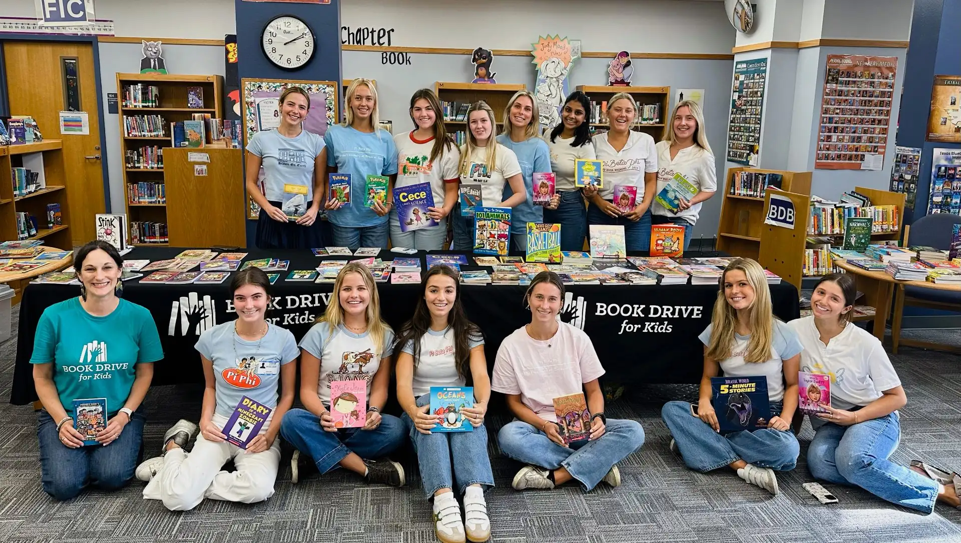 Fifteen Pi Beta Phi volunteers from UT Austin and a Book Drive for Kids team member behind a table stacked with brand-new books in a school library